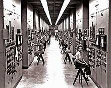 A long, high hallway, with bulky calutrons covered in switches and dials rising from the floor to the ceiling. Women sit on high chairs operating the machinery.