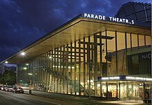 An outside, night-time shot of the building. It is well lit and has almost full-length glass panels. At the right side is the name of the drama school and also the lettering for Parade Theatres. An interior stairway is visible on the left side and a rounded wooden structure at right.