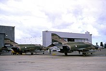 Colour photo of two military jet fighters painted in a camouflage pattern parked in front of a large white building