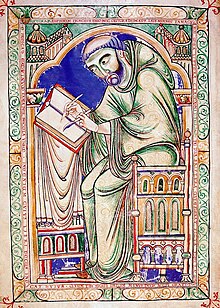 A man with a tonsure haircut in medieval robes sits at a table writing into a book with a quill.