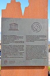 Frontale Farbfotografie einer Eisentafel mit den Logos von UNESCO und Welterbe mit einem zweispaltigen Text darunter. Die Tafel hängt an einem braunen Stein, der oben unregelmäßig gemeißelt wurde.