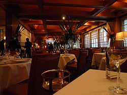 table-height view of white tablecloths, windows at right, empty glassware, dark with warm lighting