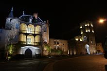 Night shot of the façade of the Moutier abbey dwelling. The square building, flanked by corner towers, has 3 storeys and a slate roof. The facade opens on the ground floor with a stone ogival door in the center, while the 2 upper floors feature a single window and wooden balconies running from one tower to the next, whose lighting creates a warm yellow hue that contrasts with the white hue of the first floor. To the right of the main building, a smaller, recessed building links the abbot's lodgings to the convent buildings, whose bell tower can also be seen in the light.