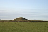 A small round hill lies beyond a field of tall grasses. The hill is also grass covered. A small entrance way to a passage into the hill is just visible at centre and a small human figure stands on top of the elevation.