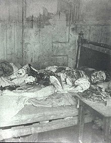 Black and white photograph of an eviscerated human body lying on a bed. The face is mutilated.