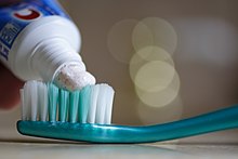 Photo with toothpaste from a tube of toothpaste being applied to the bristles of a toothbrush in the foreground