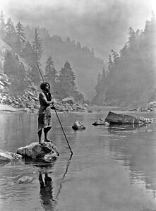 Hupa man with spear, standing on rock midstream, in background, fog partially obscures trees on mountainsides.