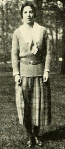 A young woman with dark hair, standing outdoors; she is wearing a blouse with a white collar under a belted sweater, with a pleated checked skirt