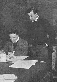 two middle-aged white men, both clean-shaven with full heads of neatly cut dark hair, the one on the left seated, the other standing