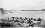Native people sit in canoes along the shore of a very wide flat river.