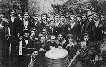 Rows of men in the woods holding musical instruments. They dress in Western or traditional styles.