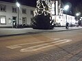 Bahnhofsvorplatz in Marburg mit weißer langgestreckte 20 als Nummer aufgemalt auf dem asphaltierten Straßenbelag. Haupteingang am Abend zur Weihnachtszeit mit beleuchtetem Weihnachtsbaum (27. Dezember 2016). Der mit einem Bordstein abgegrenzte Bereich unmittelbar vor der Treppe zum Eingang des Marburger Hauptbahnhofs (links) ist den Fußgängern und schiebenden Radlern vorbehalten, eine Nullabsenkung im Bordstein (Bordsteinabsenkung) erleichtert auch Rollstuhlfahrern den Zugang, mit einem hellen, tastbaren Aufmerksamkeitsfeld dort am Straßenrand „sehen“ auch Blinde den Beginn der Straße; die Fläche am Busbahnhof kann neben den Stadtbussen auch von Radverkehr befahren werden, die weiter nach links anschließende Neue Kasseler Straße ist eine wichtige Veloroute. Einige Fahrräder stehen an der Hauswand geparkt hinter dem Weihnachtsbaum nahe des hell erleuchteten Eingangs, nicht jedoch direkt vor der Treppe, etwas weiter links außerhalb des Bildes gibt es noch eine überdachte Abstellanlage für Fahrräder.