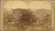 Two identical photos of a street, crowded with various horse-drawn wagons, with large storage tanks on either side and oil derricks visible in the distance. The images are mounted side-by-side on a card.