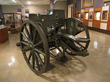 Colour photo of a green artillery gun inside the gallery of a museum. Other exhibits are visible behind the gun.