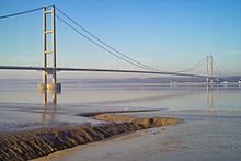 The near pier of a suspension bridge spanning calm blue waters of a wide river estuary.