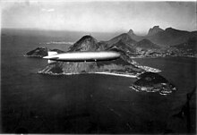 A black-and-white photograph. The Graf Zeppelin flies from left to right, with a high sun glinting off its envelope. The two right and the rear engine nacelles are visible, and the very base of the control room is catching the sun. Behind it are rocks, mountains and sea and a large city, Rio de Janeiro, built around a bay.