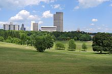 A rolling, grassy landscape with some trees beyond which some tall buildings can be seen under a blue sky with clouds. The tallest are light-colored modernist structures in the center of the image; smaller, older and darker buildings, including two church spires, rise above the treetops closer to the edges.