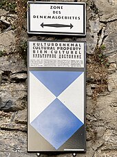 Ein Blechschild, das das Schutzzeichen für Kulturdenkmäler und einen Erläuterungstext zeigt.