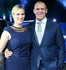 A man and a woman side by side, smiling and smartly dressed, she in dark blue sleeveless dress and he in dark blue lounge suit