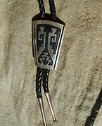 silver overlay bolo tie, by Tommy Singer, c. 1980s. This is an example of a Navajo copy of Hopi silver overlay technique, evident from the absence of matting on the black oxidized surfaces of the bottom silver sheet, or small, repeated, closely packed chisel strokes, very taxing on the silversmith, especially the eyes.[59]