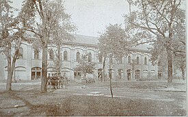 Black and white photograph of a long building, with trees and a group of soldiers
