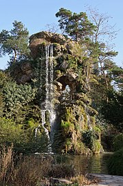 Frontale Farbfotografie eines moosbedeckten Felsens, auf dem Sträucher und Bäume wachsen. Ein kleiner Wasserfall fließt vom oberen Felsen in den Teich am unteren Bildrand, der von Schilfen umgeben ist.