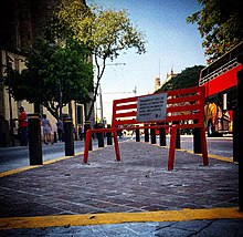 A red bench with a plaque that reads "In memory of all the women murdered by those who claimed to love them or just because they were women" in Spanish.