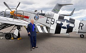 Olsen, using a cane for support, standing in front of a P-51 Mustang on static display. The airplane is painted in Invasion stripes.