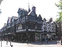 A complex range of predominately black-and-white timber-framed buildings seen from a corner. To the left is a range of shops with an arcade at ground level and windows and decorated gables above. The building on the corner has four gables of differing size and height and a spire; this building has a painted effigy in the corner of the middle storey. To the right is a simpler building; it is a public house in three storeys with three dormers.