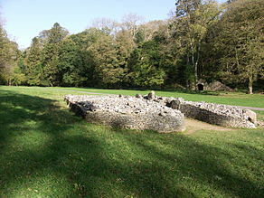 Front view of cairn, from its right side, its boulders retained by a short wall that forms a courtyard at its entrance. The cromlech is set in flat ground of short grass (in dappled sunlight in the foreground and full sun elsewhere), dissected by a path passing behind it. Trees are mainly in leaf to its rear, among which a limestone kiln is visible at the foot of the gorge.