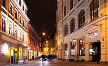 Nachtaufnahme von einer beleuchteten Straße mit alten Häusern. In den Erdgeschossen sind Läden und Gastronomiebetriebe. Ein Auto und Menschen laufen verschwommen auf der Straße.