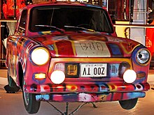 An upside-down image of a car hanging from a ceiling with brightly coloured artwork painted on the exterior. The license plate reads ZOO TV.