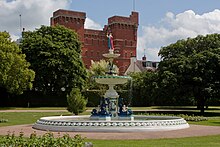 Ornamental fountain in circular pool surrounded by grassy areas. In the background is a red brick building.
