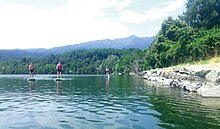 Paddleboarders are on a green-blue lake with some small ducks closer to the rocky bank. There are lots of trees, with mountains and blue sky behind.