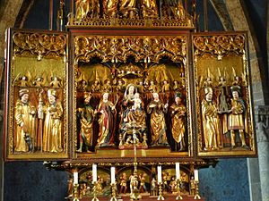 The central section of an elaborately carved, painted and gilded altarpiece showing the Virgin Mary and Christ Child seated in majesty and surrounded by saints and angels. Although the flesh and some details are painted in colour, most of the surfaces are gilt. The figures are all chubby-faced and have a charming quality.