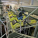 Colour photo showing a battery of five screw presses in a cooperative. Above, an endless stainless steel screw used to bring the harvest in the press. The selection of each unit is done by means of an automatic door. The winery is very clean: white walls, machines and media access in painted steel and concrete floor gallery and coated with a washable paint.