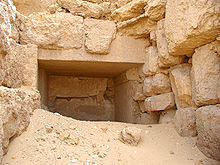 entrance of a stone lined corridor filed with desert sand amidst stone rubble