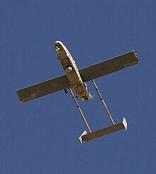 A large airborne machine photographed in flight from the ground looking up. The machine is pointed toward the top left side of the photo. Large wings can be seen protruding from the vehicle, along with the tail fin and metal peinces that attach it to the body of the aircraft. Visible in the machine's underbelly are a camera and landing gear, tail hook, and a blur in the back where a small propeller responsible for powering the machine can be found.