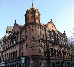 Photograph of a brick building with gables, archways, an octagonal corner tower, and a four-faced clock.
