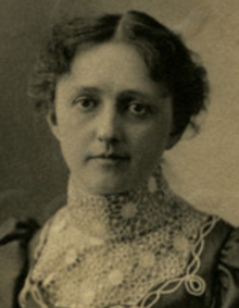 A sepia-toned photograph of a white woman with dark hair, parted center and curling on the sides; she is wearing a high-collared blouse with elaborate lace and embroidery in the yoke