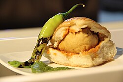 A plate of vada pav with seasoning of red chilli powder and a green chilli.