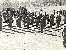 A black and white photograph of soldiers on an unsealed parade ground in a rural setting, wearing battledress uniforms, with slouch hats, webbing and rifles.