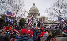 A large crowd stands outside the Capitol building while waving American flags and wearing merchandise expressing support for Donald Trump.