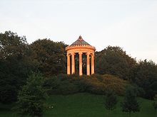Farbfotografie in der Untersicht eines Rundtempels mit Säulen und einer goldenen Kugel als Spitze. Der Tempel steht auf einem Hügel mit Büschen und ist von Bäumen umgeben.
