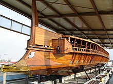 A galley sitting on struts on land under in an open-sided shed seen from the front; it has a large cast bronze ram along the waterline and an eye of Horus painted above the ram.