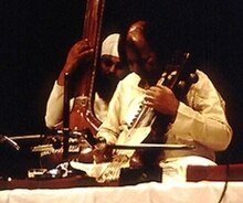 Zwei Männer sitzen auf einer Platform, einer spielt ein Streichinstrument und ein anderer zupf eine langhalsige Laute im Hintergrund.