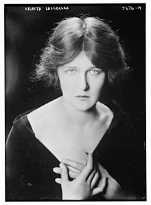A white woman with light eyes, wearing a dark garment with a deep V neckline, hands clasped to her chest