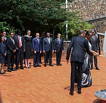 Rechts im Bild halten Paul und Jeannette Kagame wohl eine Fackel in eine Richtung, die nicht mehr im Bild ist. In wenigen Metern Abstand hinter ihnen stehen eine Reihe Personen in dunklen Anzügen und Kleidern und sehen ihnen zu.