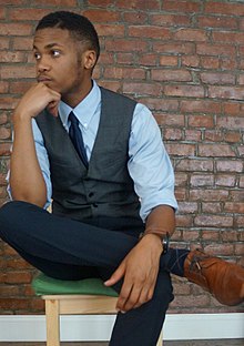 A young Black man dressed in vest and tie, with leg crossed over his knee and his chin resting on his hand. He looks off-camera to the left.