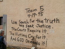 A woman, at the left of the image, is reading a black spray paint message written on a brick wall. The message reads "Team 5 4–19–95 We Search For the truth We Seek Justice. The Courts Require it. The Victims Cry for it. And God Demands it!"